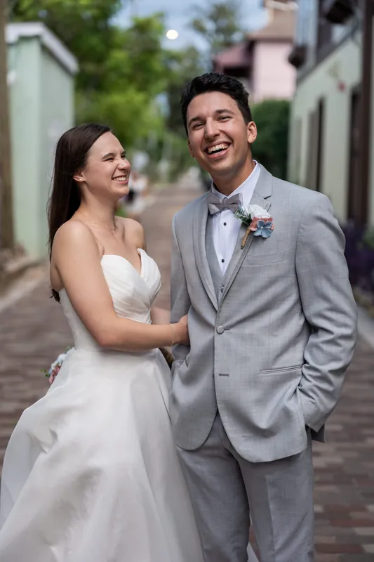 Two people laugh together on a brick street lined with greenery, one in a white dress and the other in a gray suit.