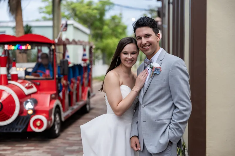 The pair poses near a whimsical red vintage train car at The White Room, with her holding his arm and both looking relaxed and happy.
