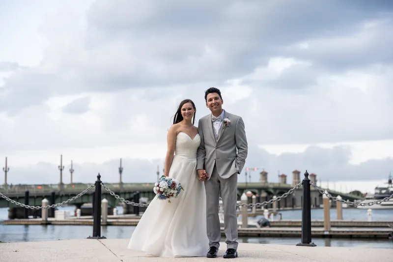 Two people stand on a rooftop holding hands, with a waterfront and historic city skyline visible behind them under a cloudy sky.
