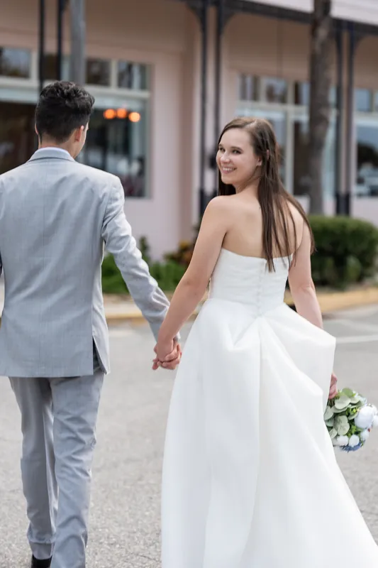 From behind, the couple walks slowly down a colonial-style street at The White Room, her train trailing behind her white gown as they hold hands.
