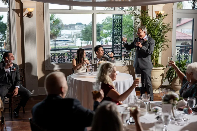A speaker stands addressing seated guests during the reception at The White Room, with waterfront views framed by tall windows behind.