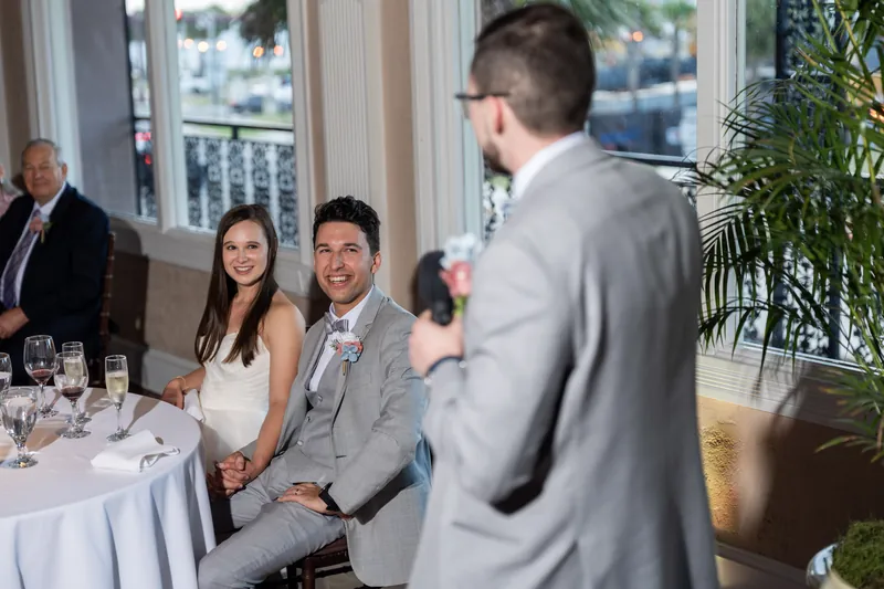 A couple seated at a table smiles while a person in a gray suit stands nearby during the reception.