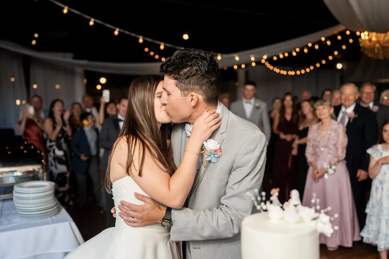 Sarah Katherine and Nathan embrace while standing beside their wedding cake under string lights.