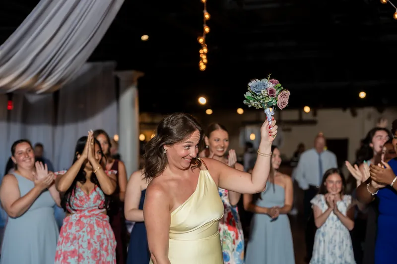 A person in a pale yellow gown holds a colorful bouquet aloft while dancing at The White Room, surrounded by guests celebrating.