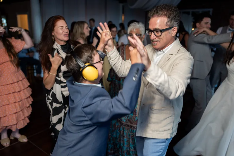 A man in a light blazer dances with a child in a navy suit as onlookers clap along at the reception.