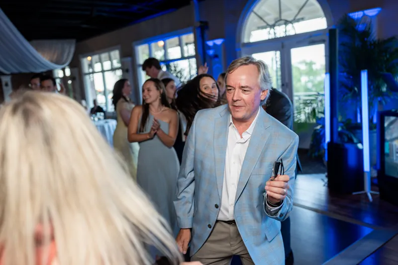 A man in a blue-gray jacket gestures while dancing near large windows overlooking the venue's grounds.