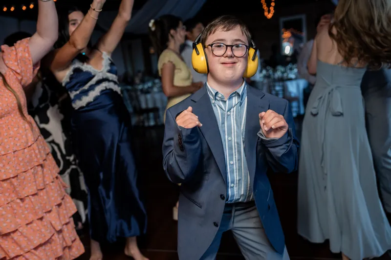A young person in a navy blazer and blue shirt dances enthusiastically among wedding guests on the lit-up floor.