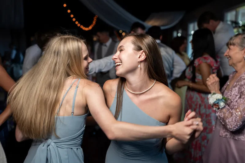 Two guests in gray dresses smile at each other while dancing, with string lights and other attendees visible behind them.