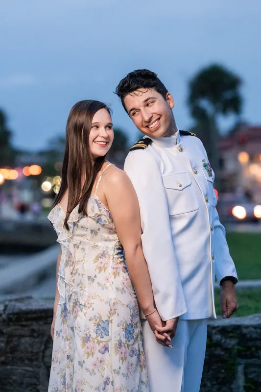At twilight, the couple stands together on the fort's grounds with lit buildings and palm trees glowing softly in the distance behind them.
