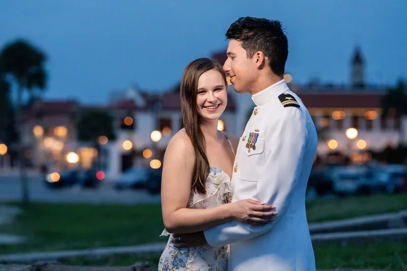 Nathan in his white naval dress uniform and Sarah Katherine in a floral gown embrace as evening lights twinkle across the waterfront beyond them.