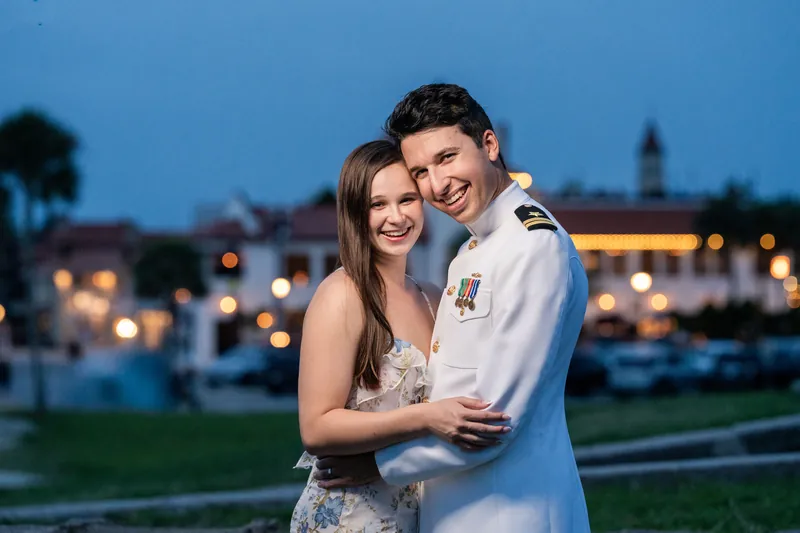 The pair smile closely together during blue hour at the historic fort, the illuminated cityscape and water visible in the softly blurred background.
