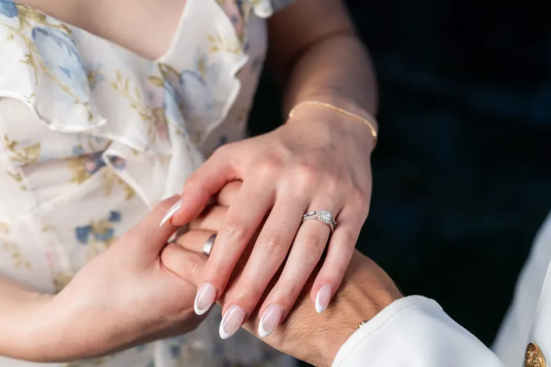 Wedding rings rest on joined hands—one adorned with a diamond band, the other a white band—against their formal wedding attire.