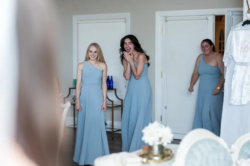 Three women in matching blue dresses stand together in a bright room with white doors and bouquets visible nearby.