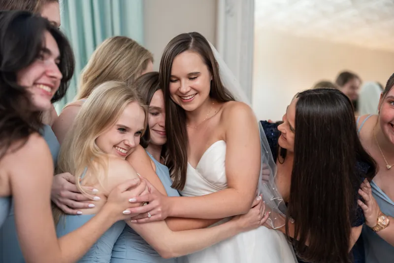 A woman in a white gown embraces several attendants in blue dresses, smiling warmly during a group moment.