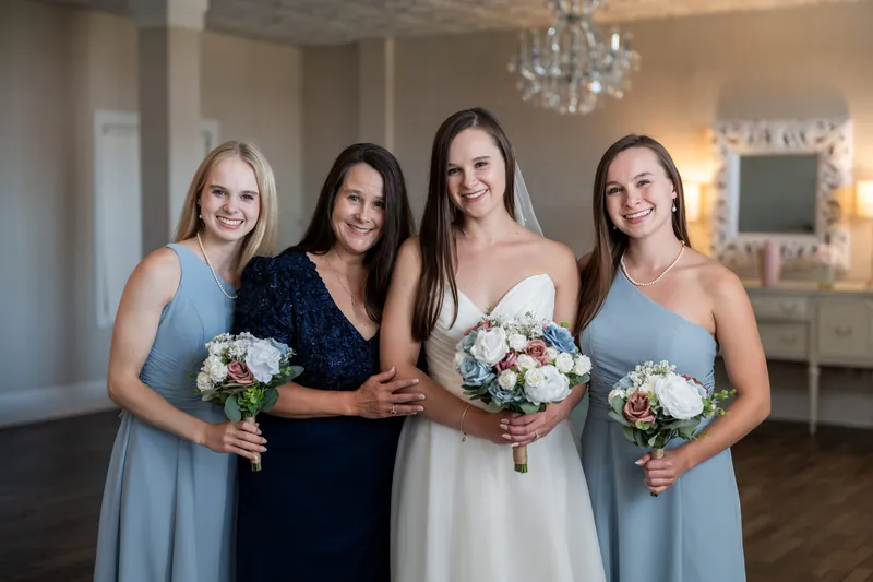 Four women hold white and green bouquets while posing together in an elegant room with chandelier and neutral furnishings.