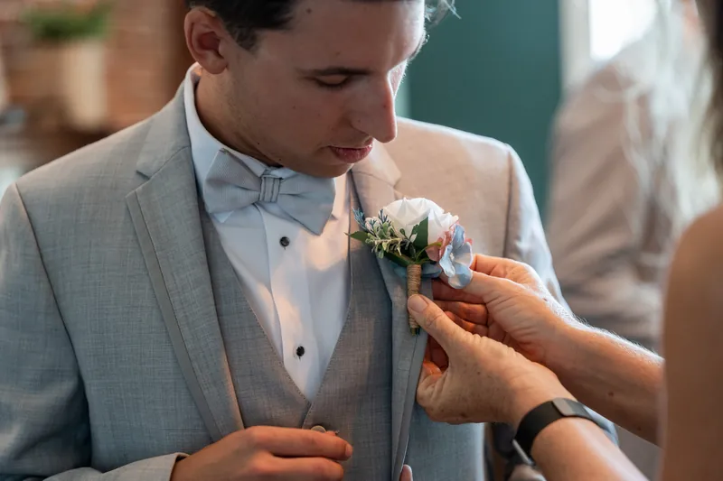 Someone in a gray suit receives a boutonniere pinned to his lapel by a guest's hands.