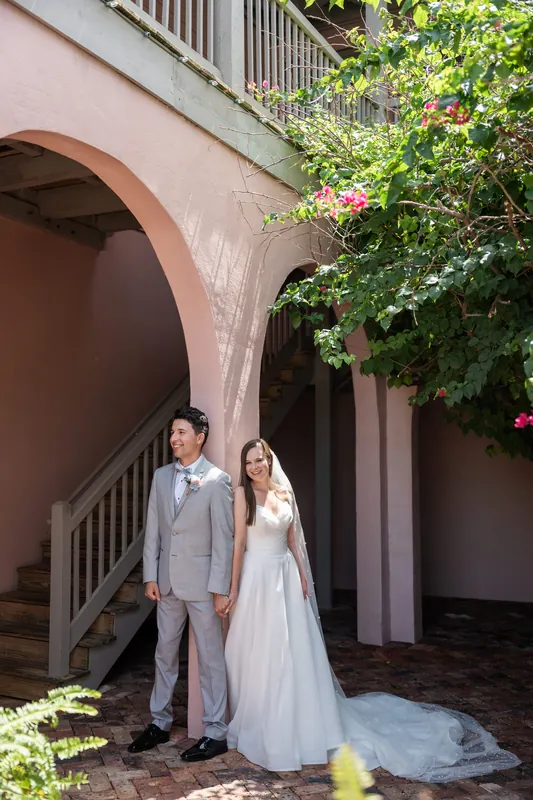 Under a curved archway with flowering vines, two people in wedding attire pose on brick flooring.