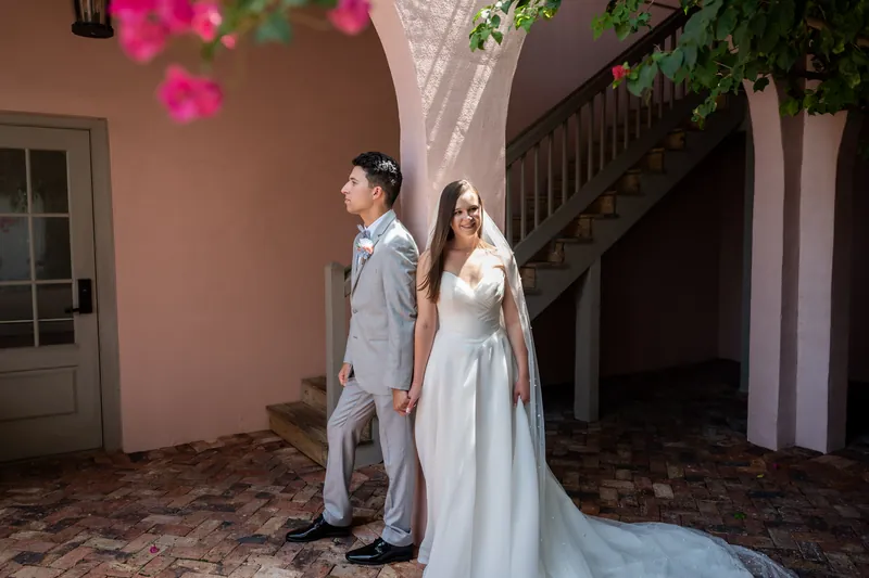 Two people stand beneath magenta bougainvillea blooms against pink stucco walls and wooden railings.