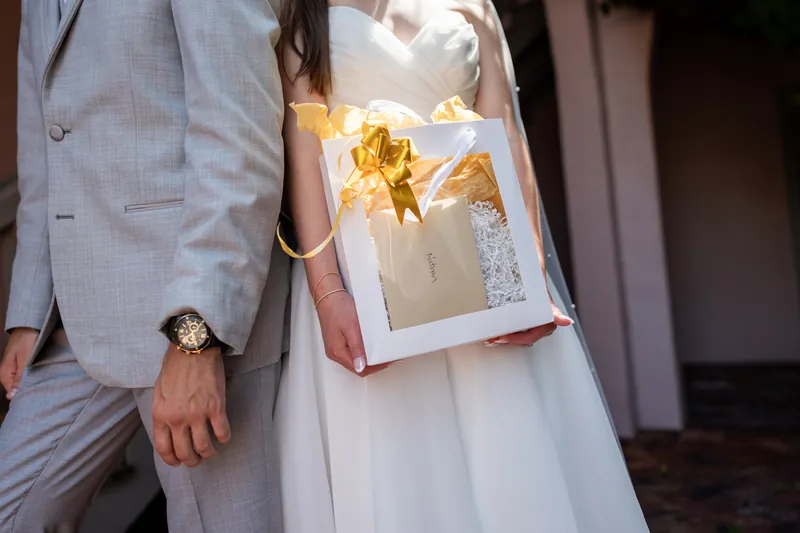 Two people in light gray suits hold a gold-wrapped box with a gold bow at The White Room, St. Augustine.