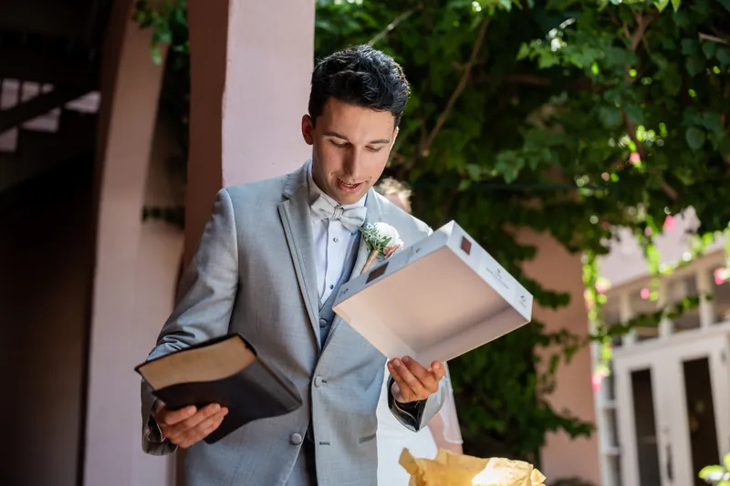 A person in a gray suit and white dress shirt holds an open gold box while standing in a courtyard with ivy-covered walls.