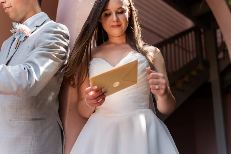 Two people in formal wedding attire examine the contents of a gold envelope in afternoon sunlight.