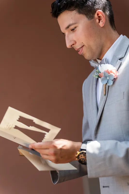 A person in a light blue shirt reads from an open card or note, wearing a boutonniere.