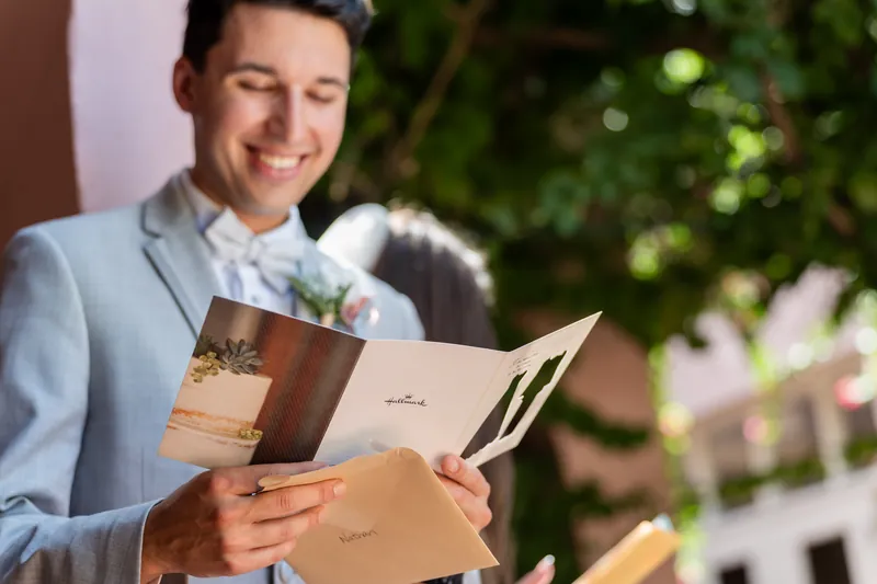Someone in a light blue suit holds open wedding programs with white flowers tucked in, smiling downward.