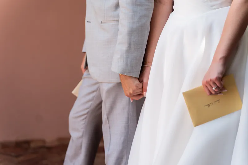 Two people in wedding attire hold hands and an envelope while standing against a textured wall at The White Room.
