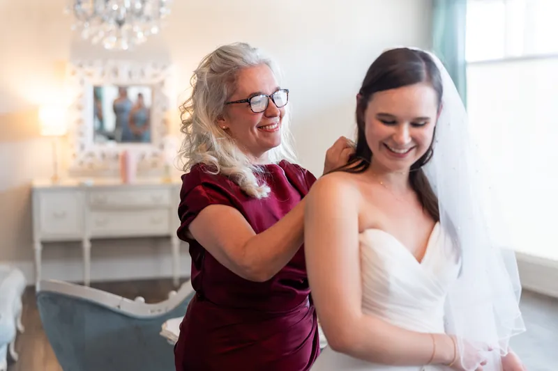 A woman in burgundy adjusts the veil of a person in a white gown inside The White Room's elegant bridal suite.