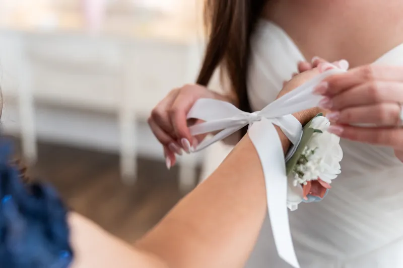 Hands fasten a delicate ribbon around a wrist corsage of white flowers at The White Room.