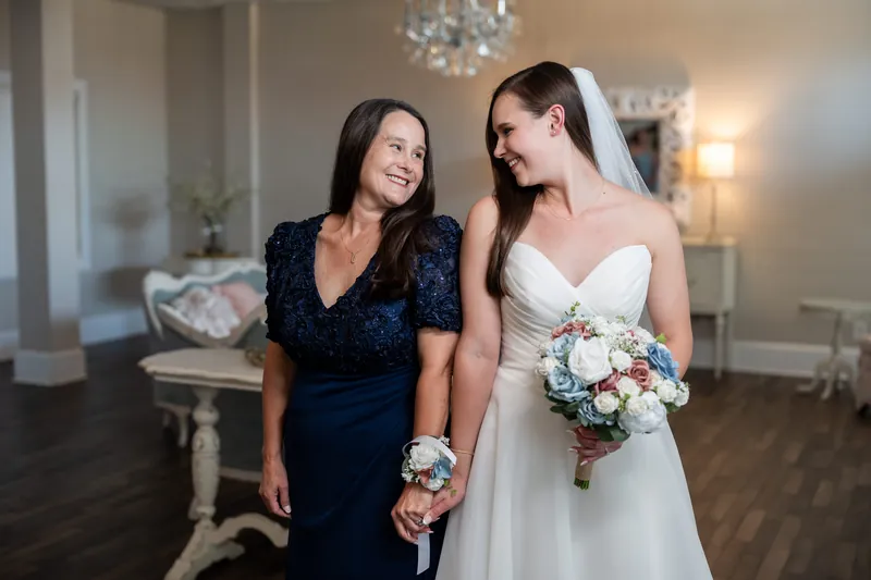 Two women stand together inside The White Room—one in navy lace holding a bouquet, the other in a sleeveless white gown with veil.