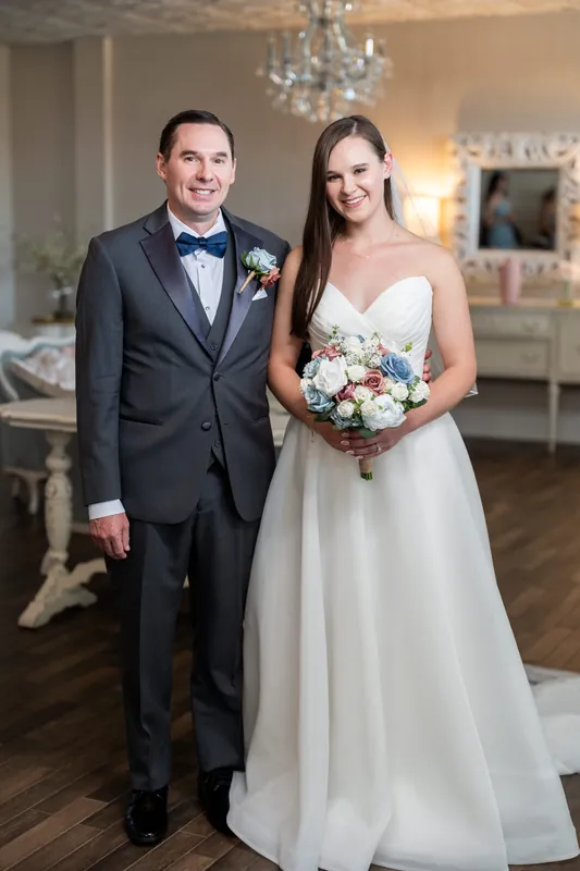 Couple in formal wedding attire in a chandelier-lit room, he in a navy suit with boutonniere, she in a strapless white gown holding a bouquet.