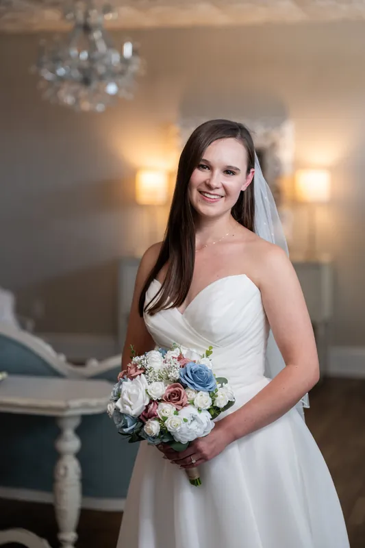 A person in a sweetheart-neckline white gown and veil smiles while holding a blush-and-blue floral bouquet at The White Room.