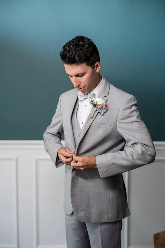Someone in a gray suit adjusts a boutonniere while looking down at The White Room.