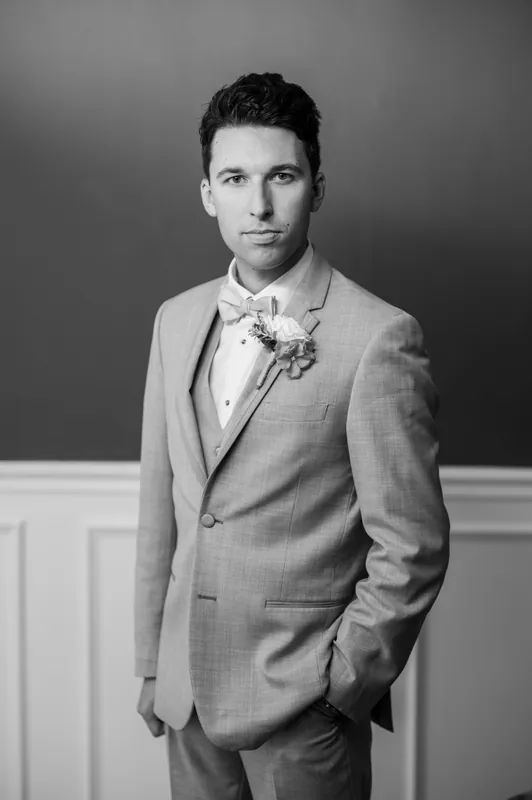 A person in a light gray suit and white dress shirt stands with hands in pockets against a neutral backdrop.