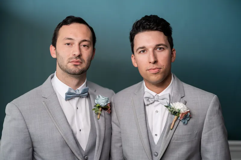 Two people in coordinating gray suits with bow ties and boutonnieres pose side by side against a teal wall.