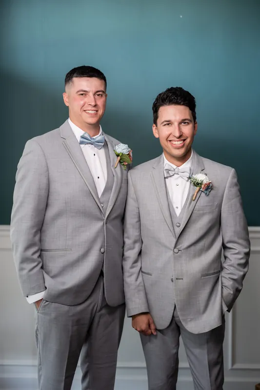 Two people in gray suits and white shirts smile together, one wearing a bow tie and the other a standard necktie, both wearing boutonnieres.