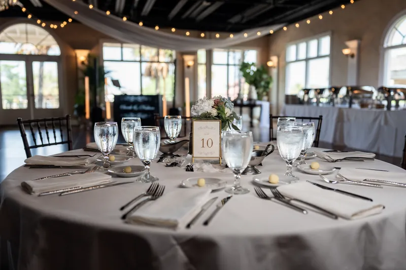 Dining table set for a reception with gray linens, numbered place cards, and water glasses casting shadows under natural light from tall windows.