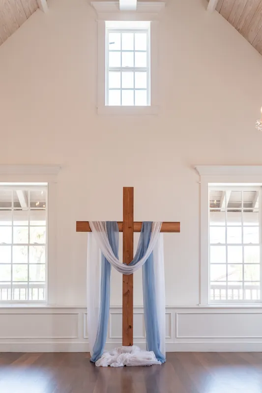 Wooden cross adorned with blue and white fabric hangs centered on a white chapel wall with symmetrical windows at The White Room.