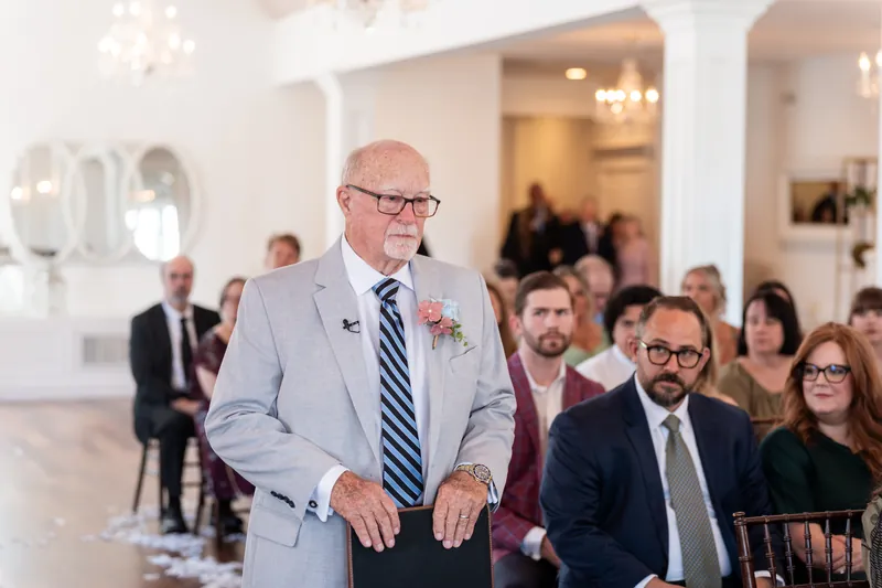 An older man in a light gray suit stands in the white interior, glancing toward guests seated behind him.