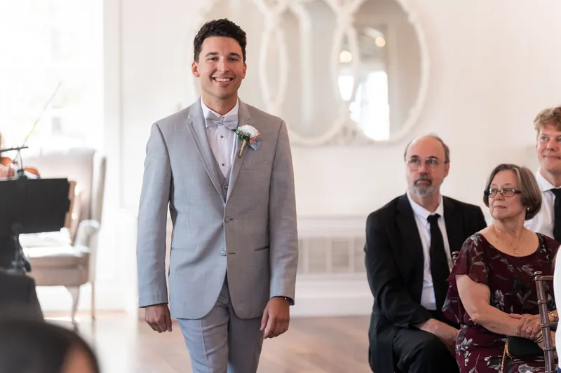 A man in a gray suit with a pink boutonniere smiles while standing near seated guests in formal wear.