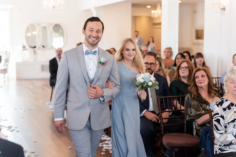 A couple walks down the aisle at The White Room—he in a gray suit, she in a blue gown holding a white bouquet—as seated guests turn to watch.
