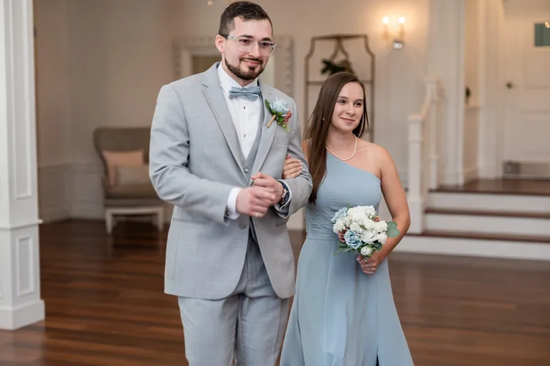 He adjusts his cufflinks while standing beside her in a pale blue dress holding a white floral bouquet in The White Room's bright, columned space.