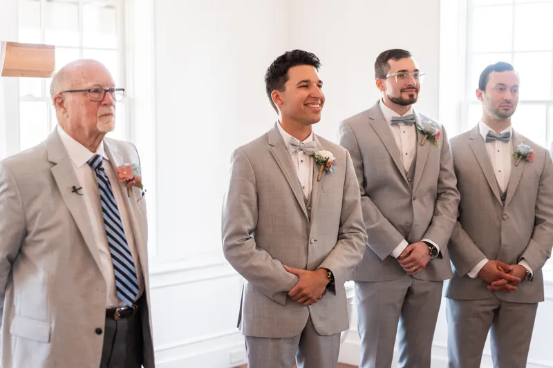 Four men in matching gray suits stand in a line at The White Room, with an older man in a lighter suit speaking to them during the ceremony.
