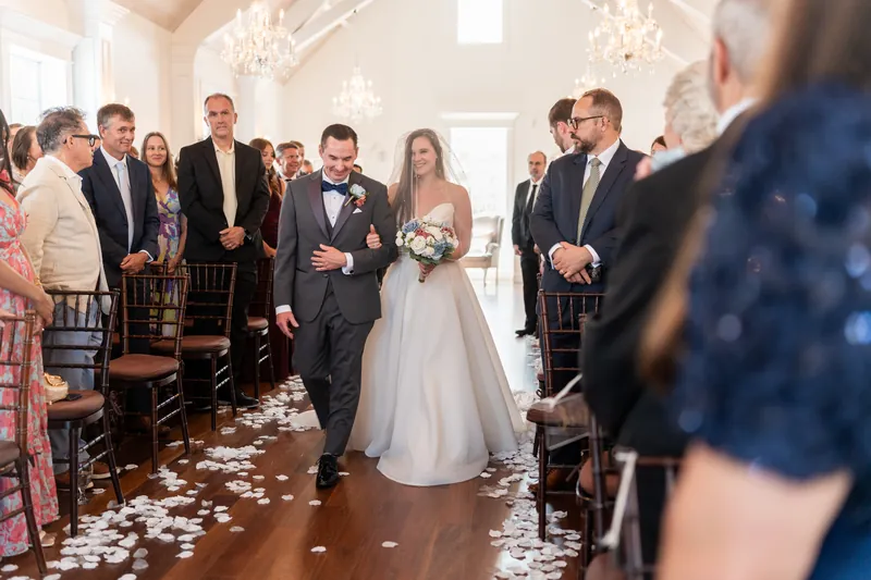 The couple walks back down the aisle after their ceremony at The White Room as standing guests turn to applaud, their faces beaming with celebration.