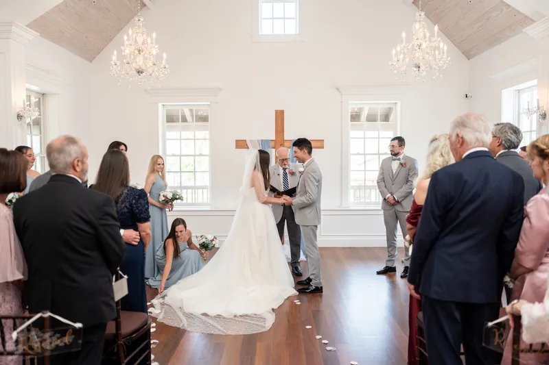 Two people exchange vows at the wooden cross while family and friends watch from the pews at The White Room.