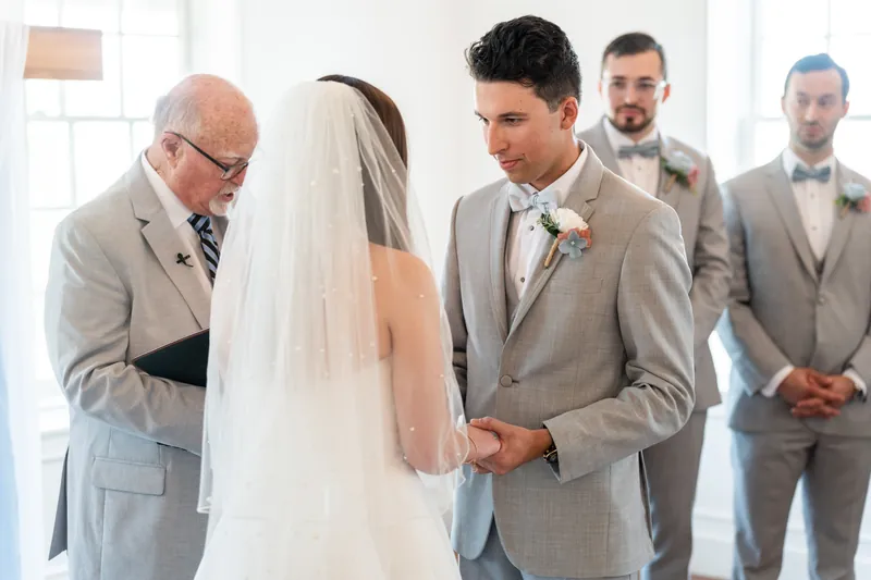 Nathan holds hands with someone in a white veil during the ceremony as groomsmen stand nearby in tan suits.