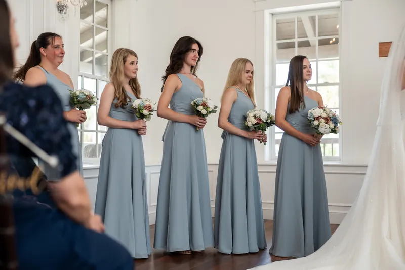 Five attendants in slate dresses hold white bouquets while standing in profile, watching a ceremony unfold nearby.