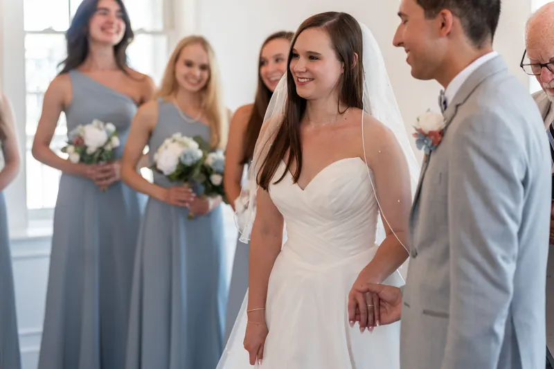 A woman in a white gown laughs with a man in a light gray suit while bridesmaids in slate dresses stand nearby at The White Room, St. Augustine.
