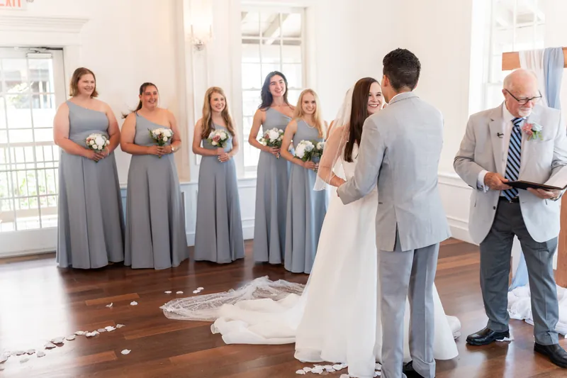 Two people embrace on a wooden floor as attendants in slate dresses and groomsmen look on at The White Room.